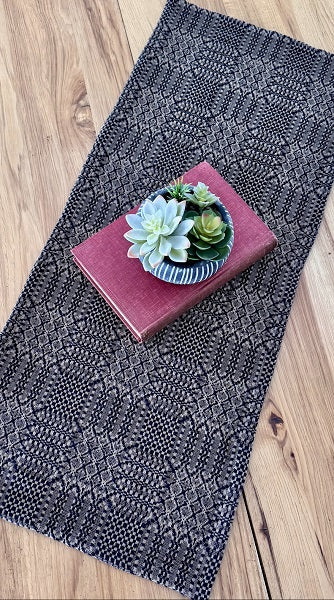 A dark gray and black textured woven placemat with a geometric pattern, styled on a light wood floor with a pink book and a small succulent plant.