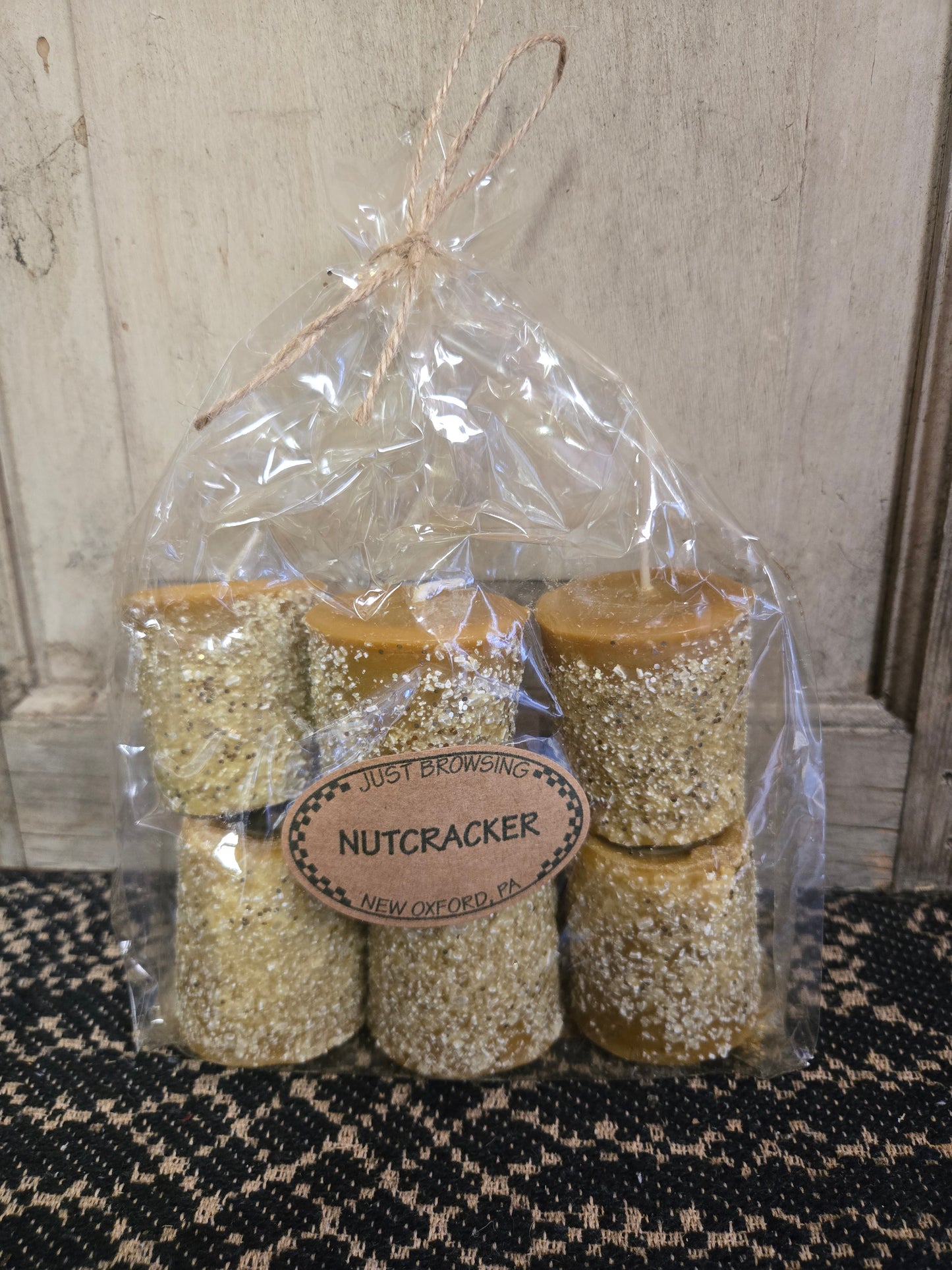 A pack of six Nutcracker themed votive candles, hand poured and hand rolled, with a beige and brown speckled appearance, packaged in clear wrapping with a brown label.