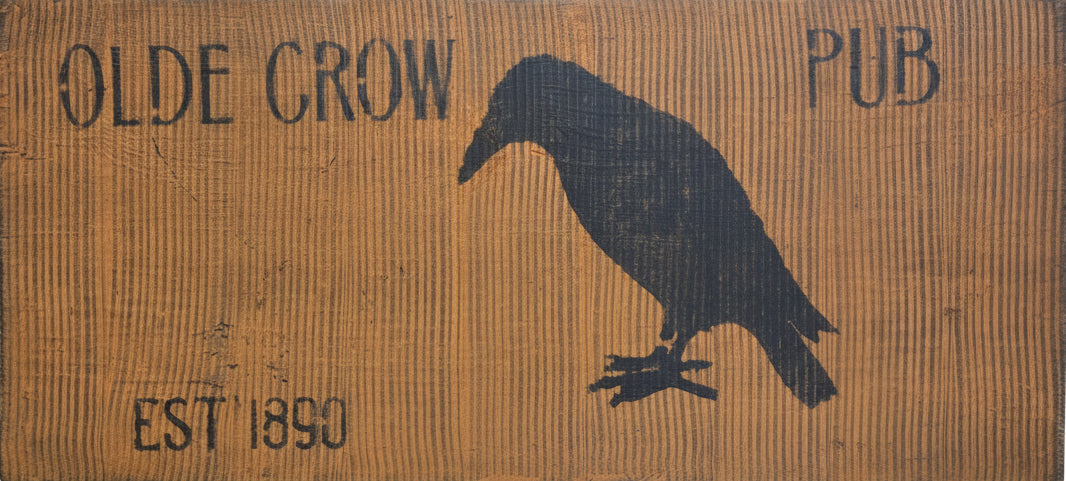 A hand-painted combed sign with 'OLDE CROW PUB' text and an illustration of a crow perched on a fence post, placed against a rustic wooden background.