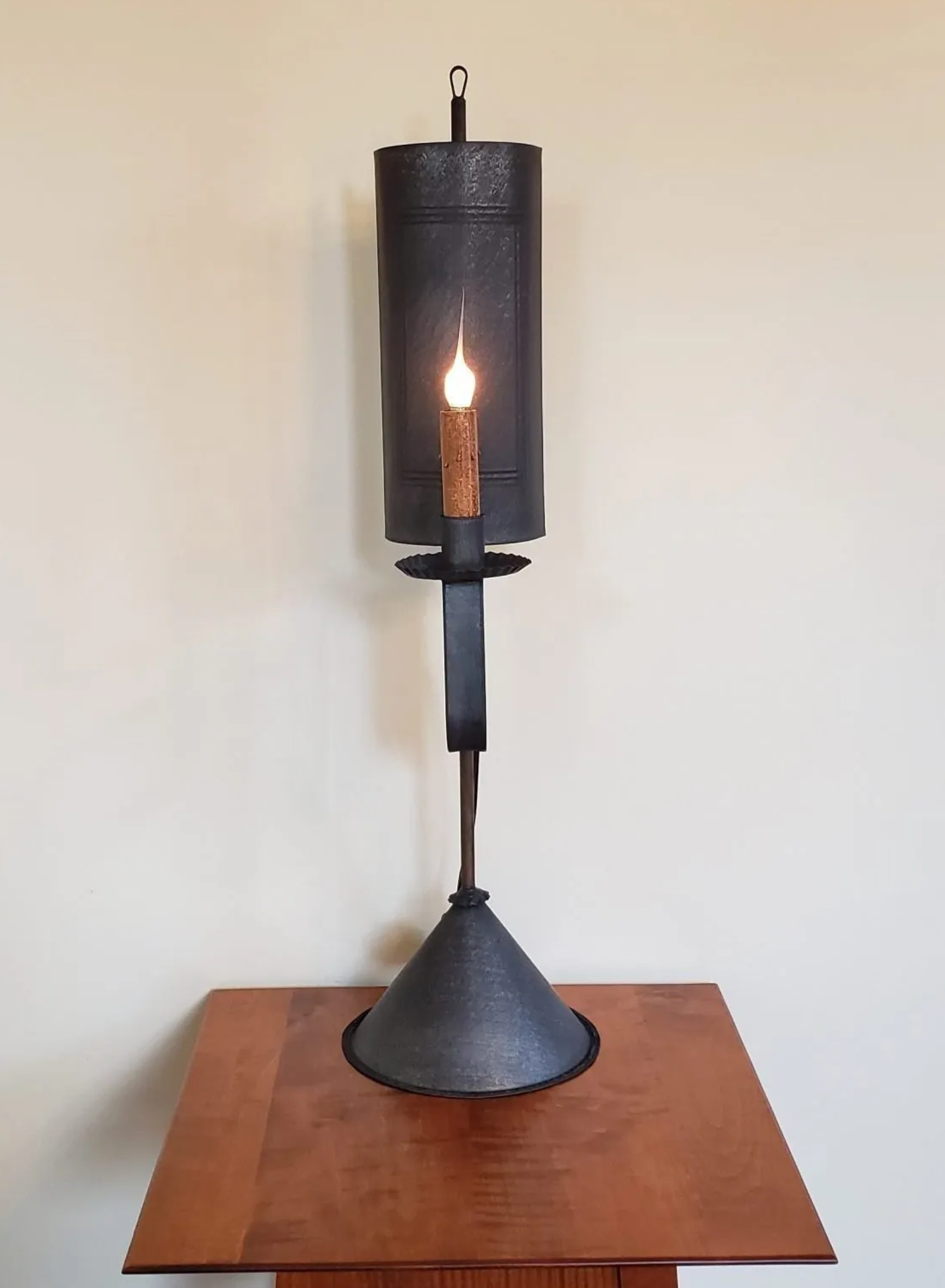 A black tin table light with a lit candle-like bulb, mounted on a wooden table against a plain wall.