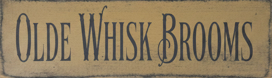 A rectangular wooden sign with the words 'OLDE WHISK BROOMS' in dark lettering, placed on a worn wooden surface.