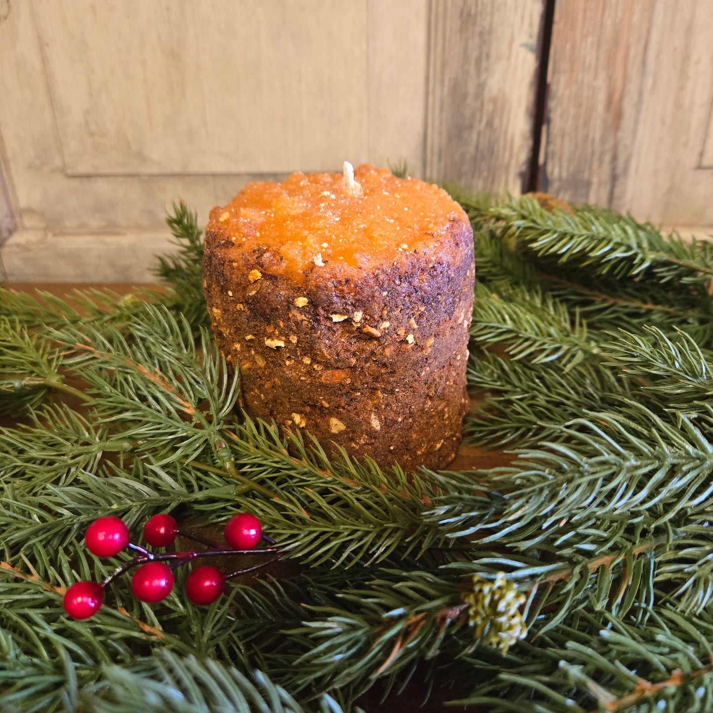 A small hearth candle with an orange tint and a brown, textured edge, set upon a bed of green pine needles with red berries.