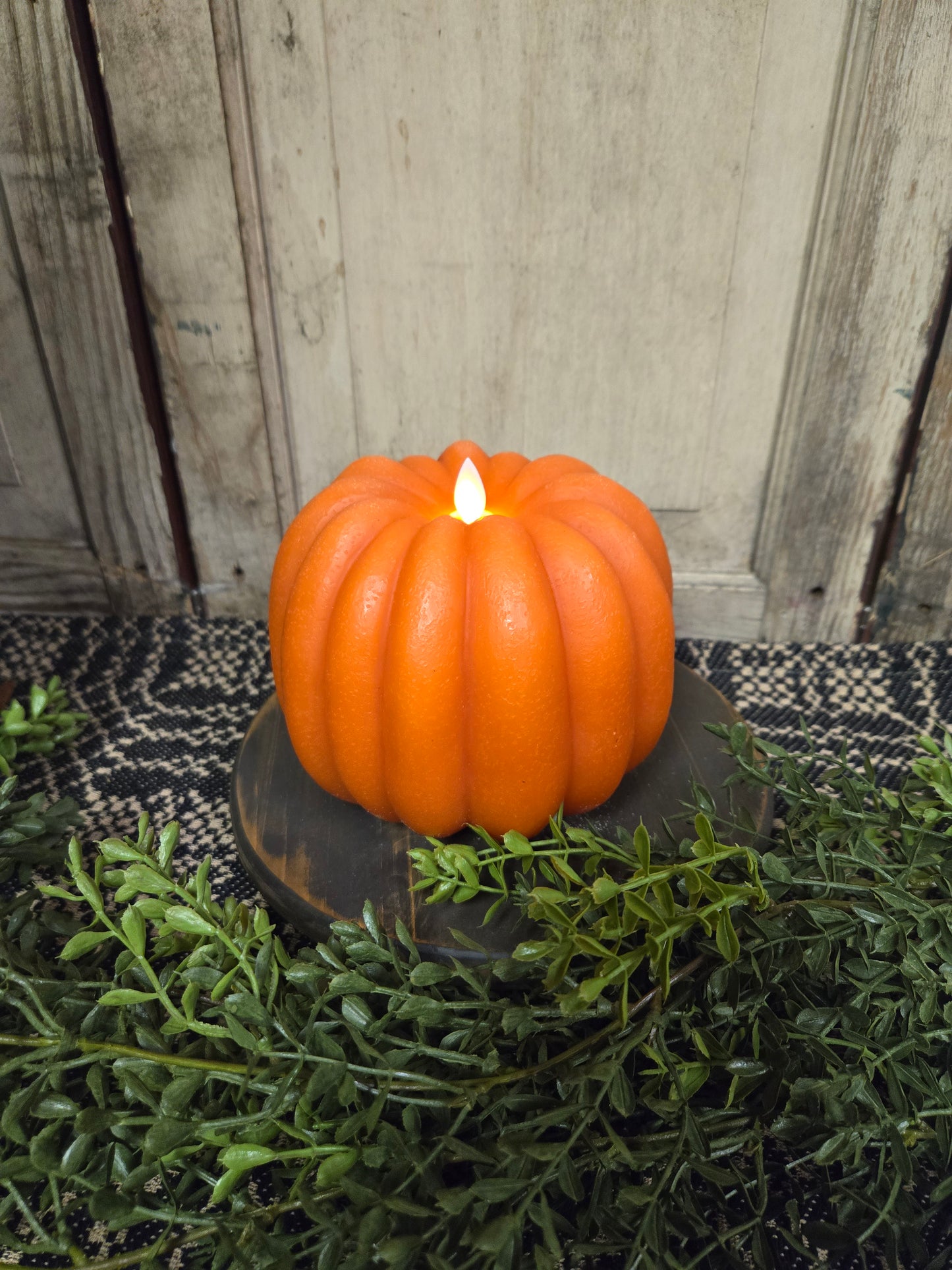 Pumpkin shaped battery operated candle on a wooden riser with greenery surrounding it