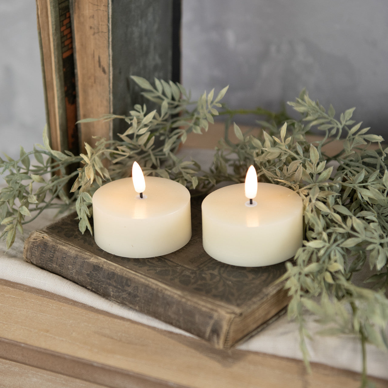Two oversized 3D flame cream tealight candles on a wooden surface with a plant wreath behind them.
