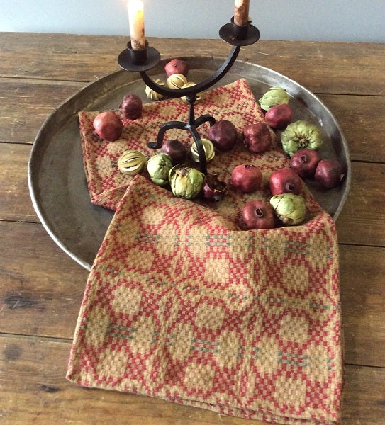 A 52" square tablecloth with a patriotic design featuring cranberry, green, and tan colors, draped over a wooden table with a decorative branch and assorted fruits on top.
