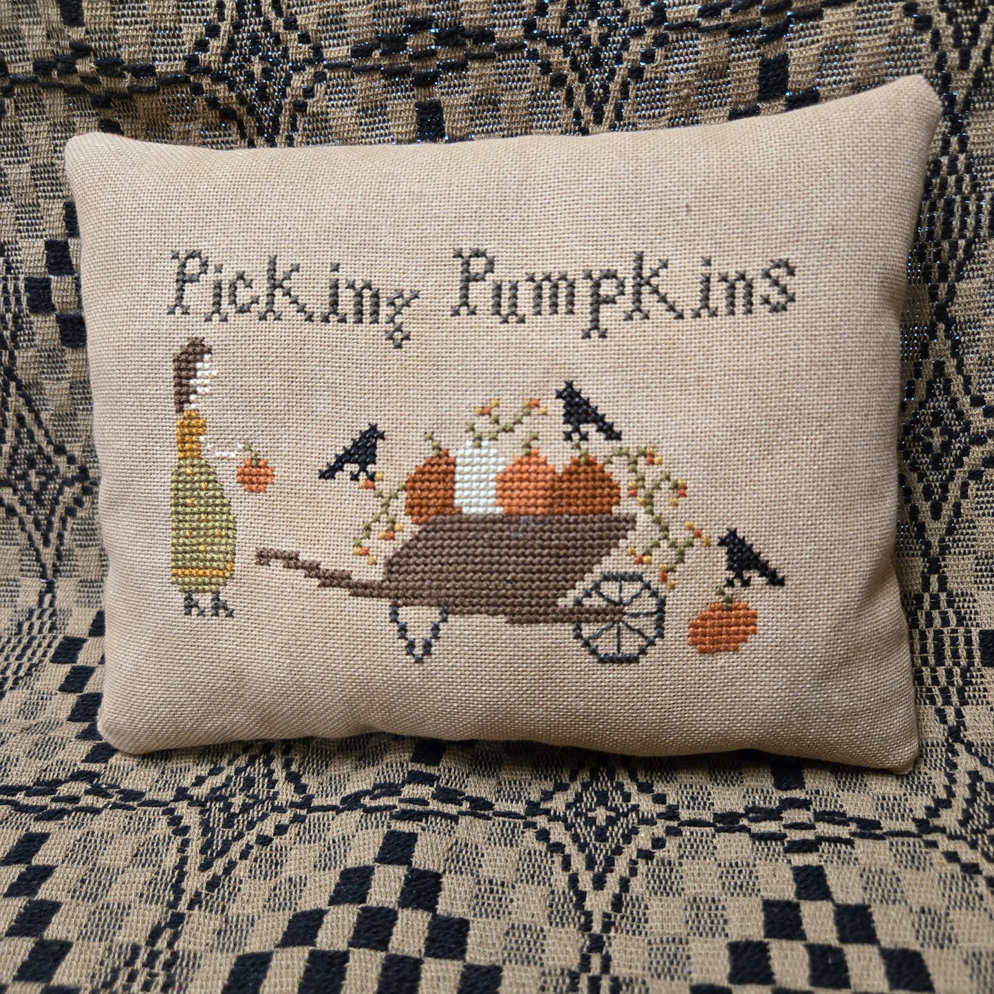 A square beige cross-stitch pillow featuring the text 'Picking Pumpkins' and a stitched illustration of a woman, pumpkins, a wheelbarrow, and birds, displayed on a patterned woven blanket.