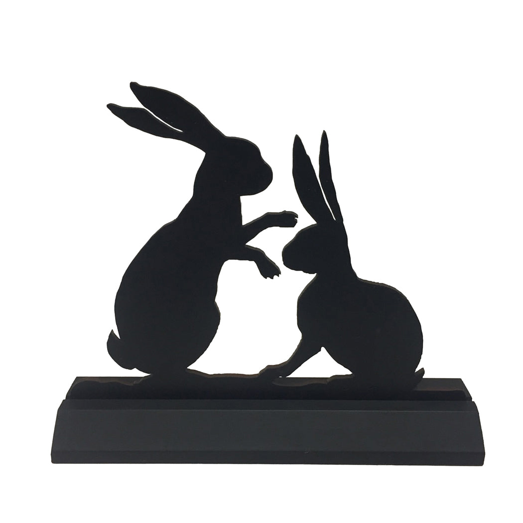 A flat black, laser-cut wood silhouette of two rabbits standing and interacting with each other on a solid wood base.