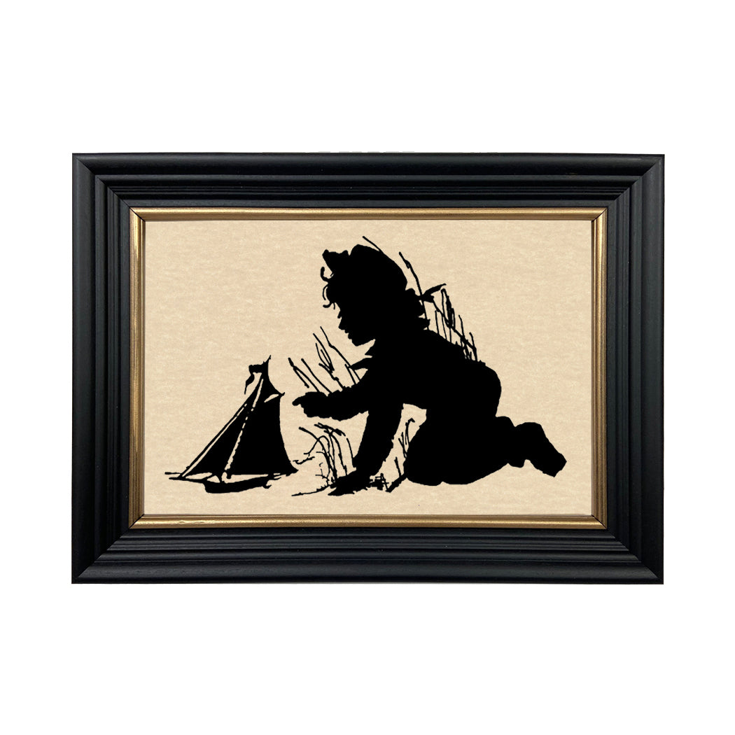 Framed paper cut silhouette of a person playing with a sailboat, predominantly black with gold trim on the frame.