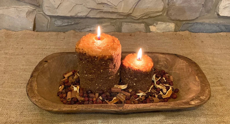Two hearth candles on a wooden platter with a lit candle, surrounded by ingredients such as cinnamon sticks and orange peels, indicative of their scent profile.