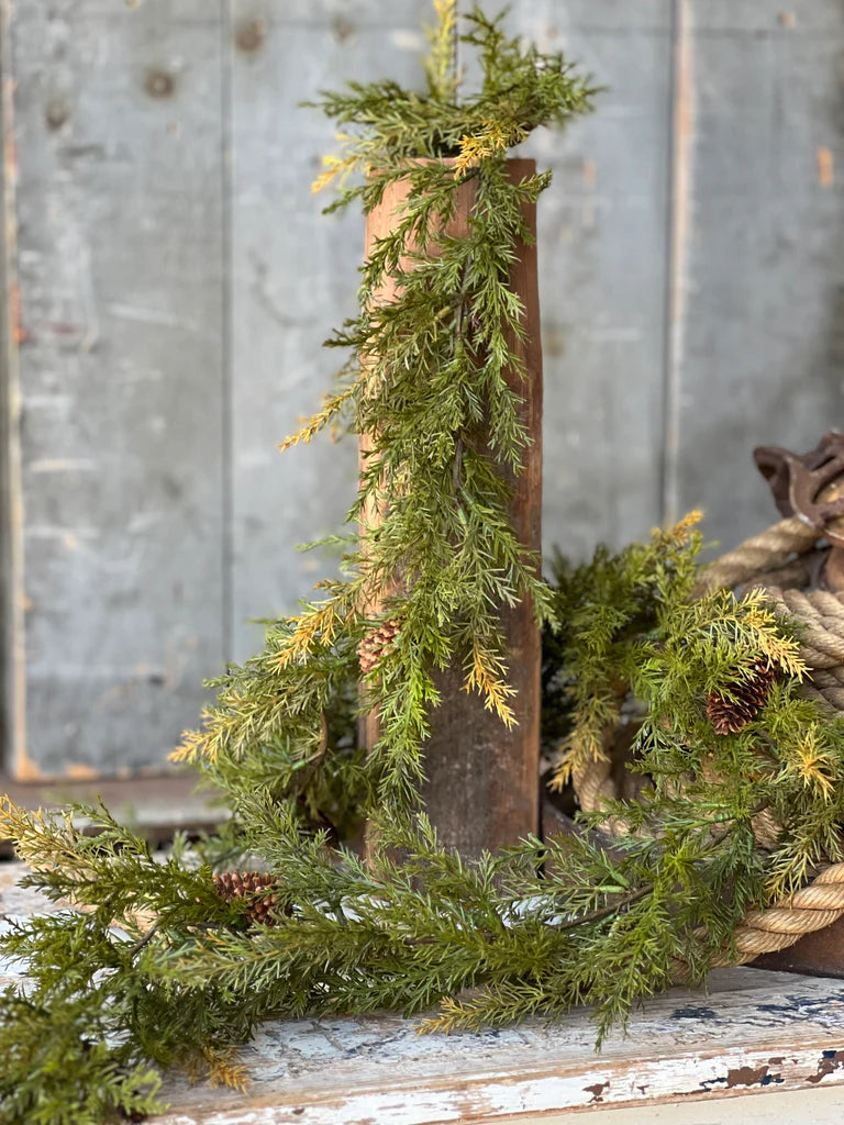 A garland made of green pine branches with brown pine cones, draped over the edge of a rustic wooden object.