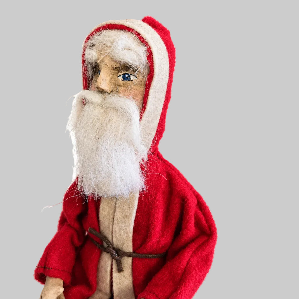 A handcrafted Primitive Belsnickel doll with a red outfit and white beard, sitting on a stool against a gray backdrop.