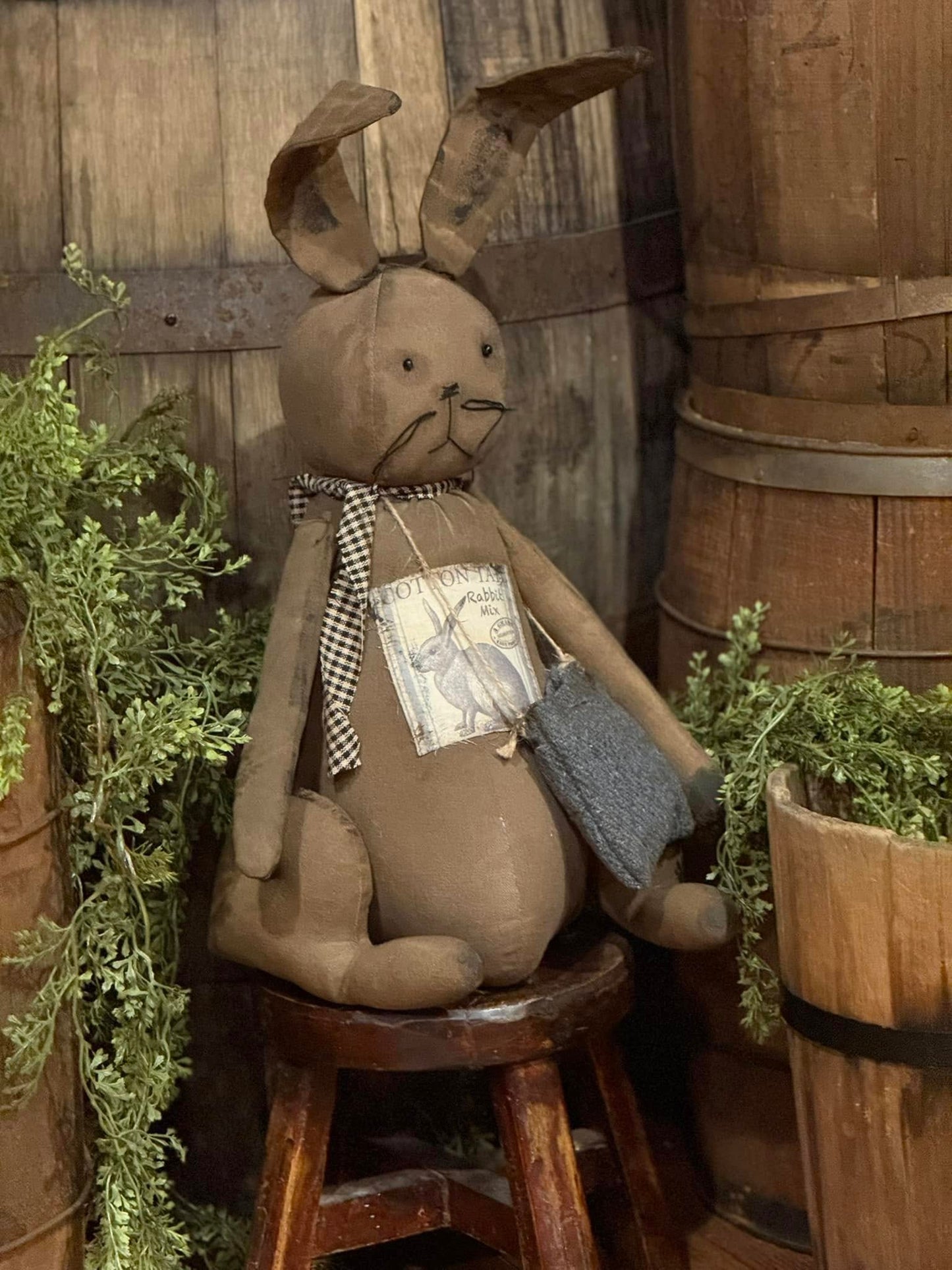 A handcrafted Primitive Cottontail Bunny doll with a brown body, beige details, and a checkered scarf, seated against a rustic background.