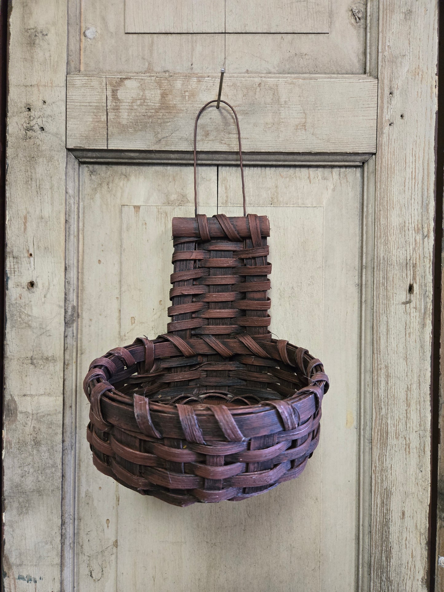A handmade Primitive Cupboard Basket with a burgundy color, hanging against a rustic wooden background.
