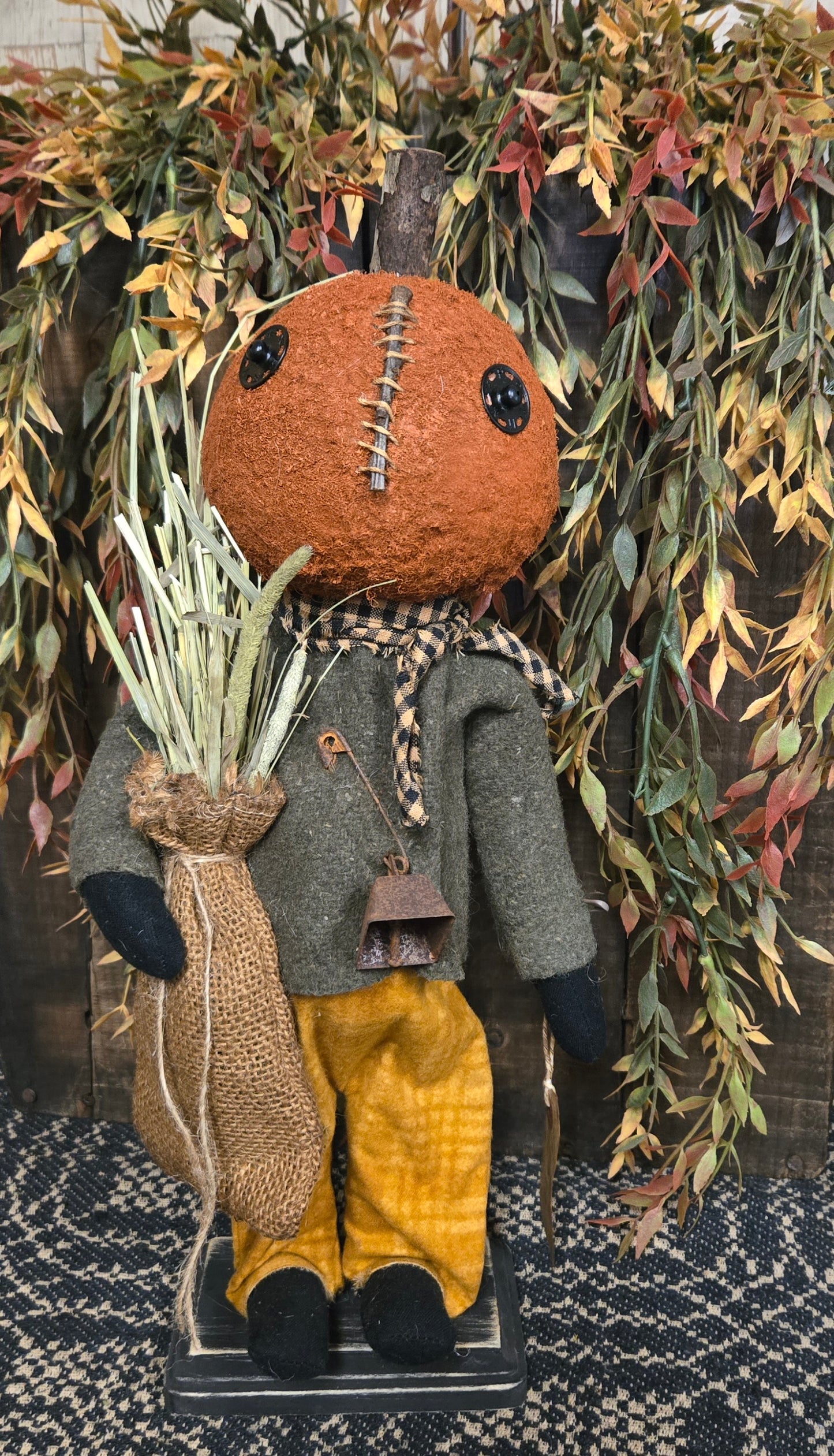 Pumpkin doll with a brown head, gray coat, yellow pants, and black shoes, surrounded by greenery.