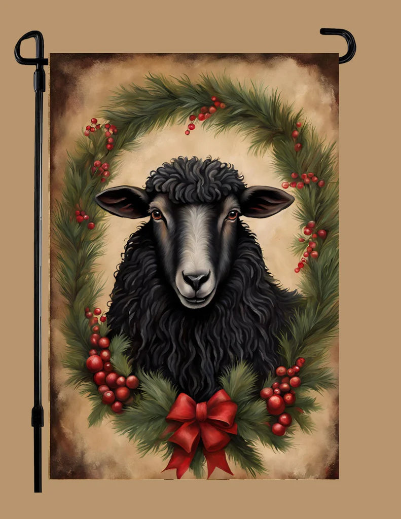 A decorative garden flag featuring a black sheep centered within a green wreath with red berries and a red bow, shown hanging on a black garden flag pole against a tan background.