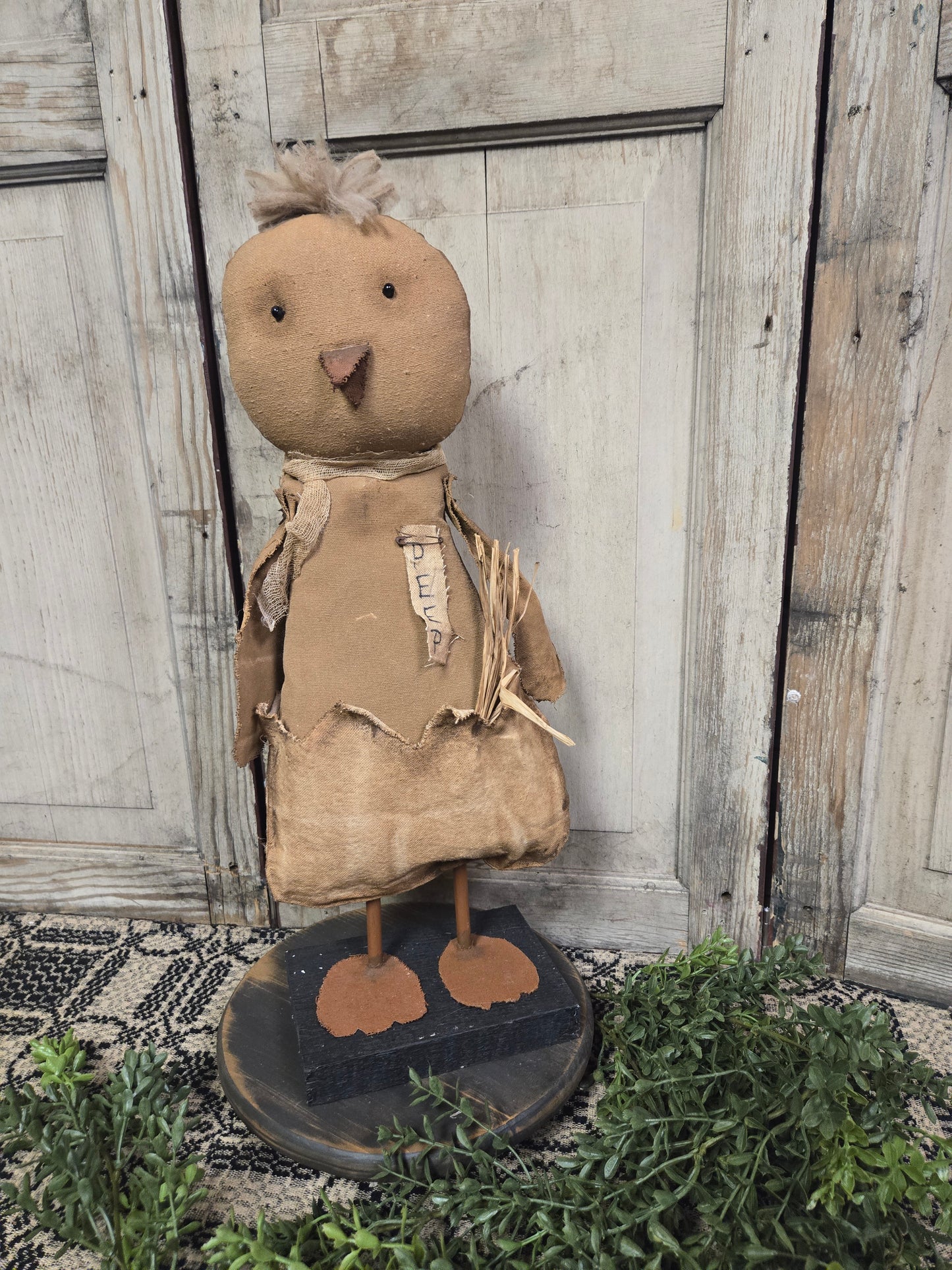 A primitive style sculpture of a chicken with a realistic design, standing against a wooden backdrop with greenery on the ground.