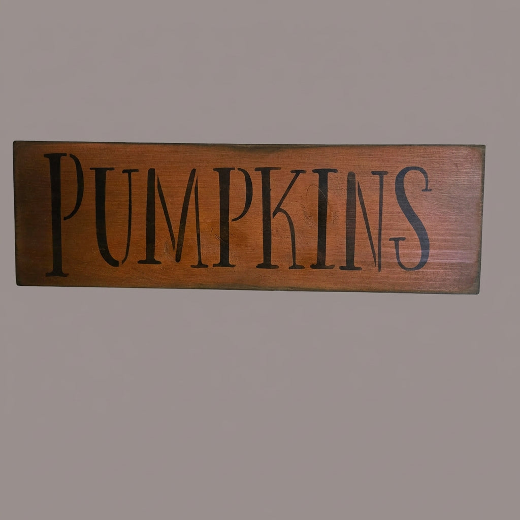 A rustic wooden sign featuring the word 'PUMPKINS' in large, black serif letters against a warm brown wood-grain background.