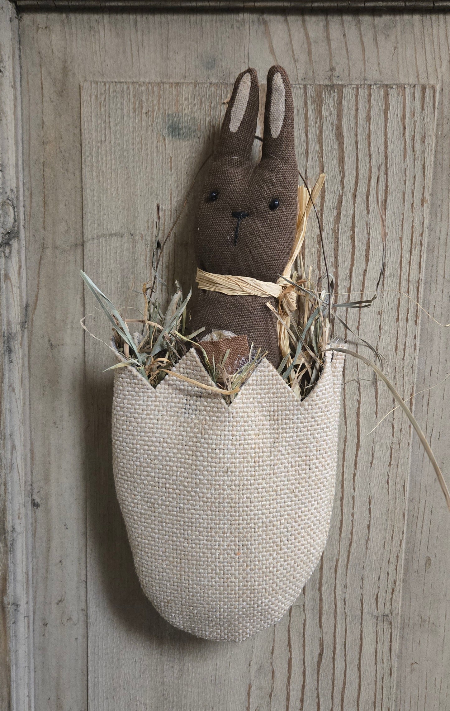 Textile rabbit figure in an egg-shaped basket on a wooden wall.