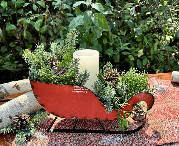 A red metal sleigh with greenery and a candle inside, displayed on a table with a nature backdrop.