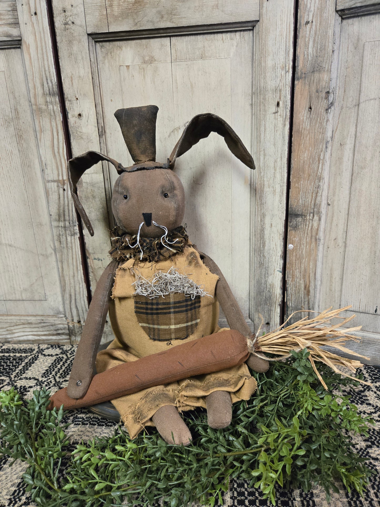 A figurine of a sitting bunny with a carrot, wearing a brown dress and a hat, positioned in front of a wooden background.