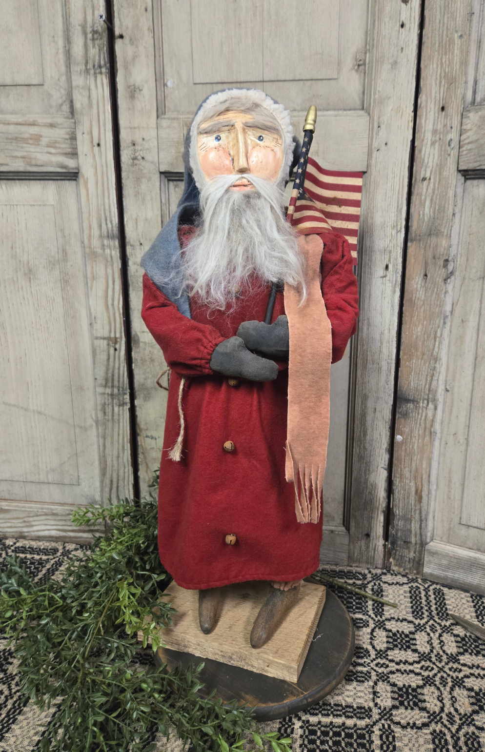 Clay faced Santa figurine holding a flag, dressed in red and gray, with a backdrop of a wooden door and foliage.