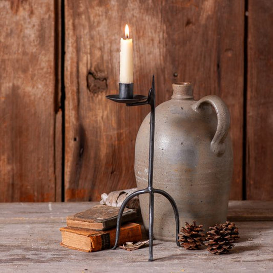 A single taper candle placed in a wrought iron candle holder with an aged finish, set on a wooden surface with a pine cone and an old book in the background.