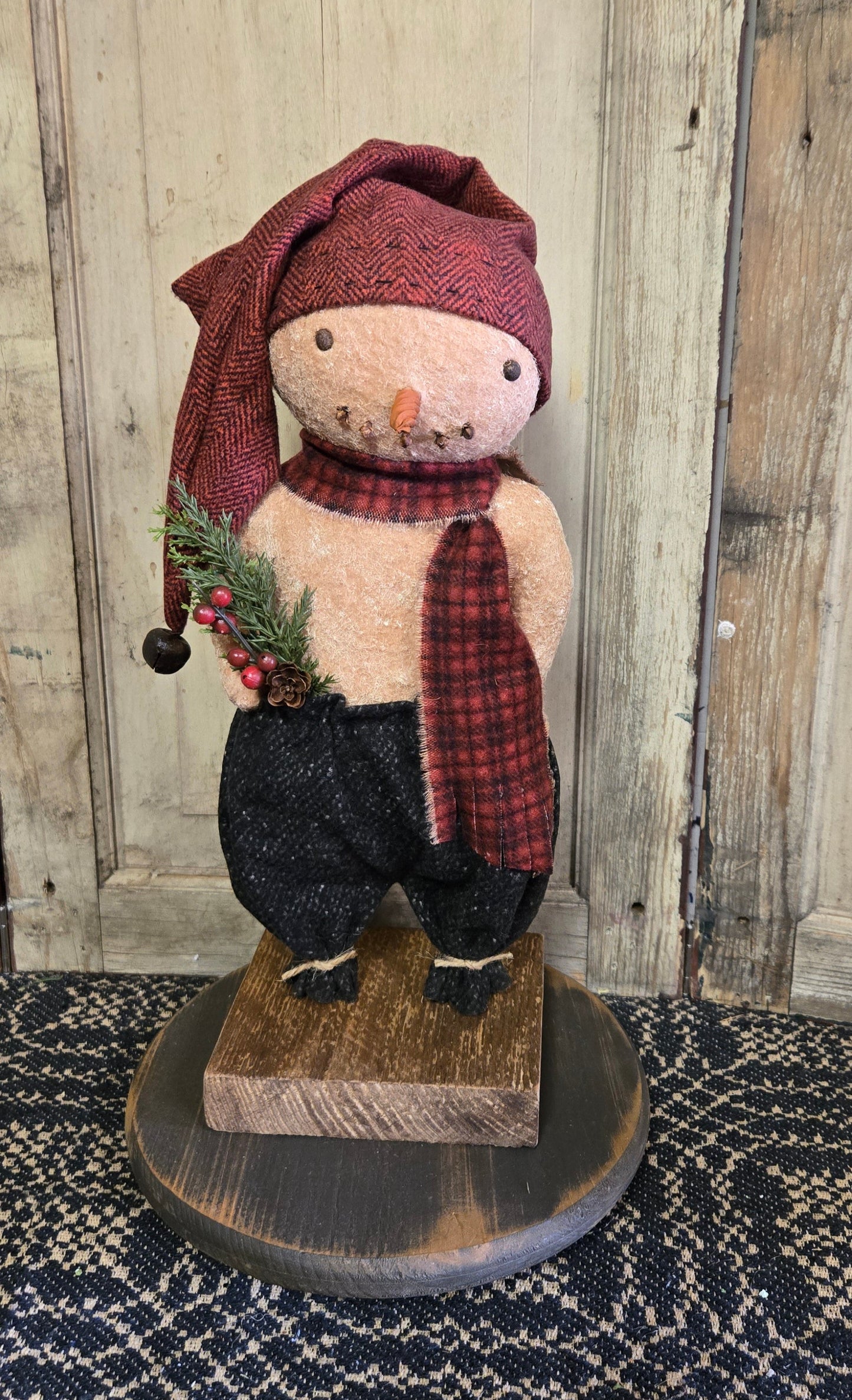 Snowman doll dressed in red and black with a sprig of pine displayed on a black wooden riser