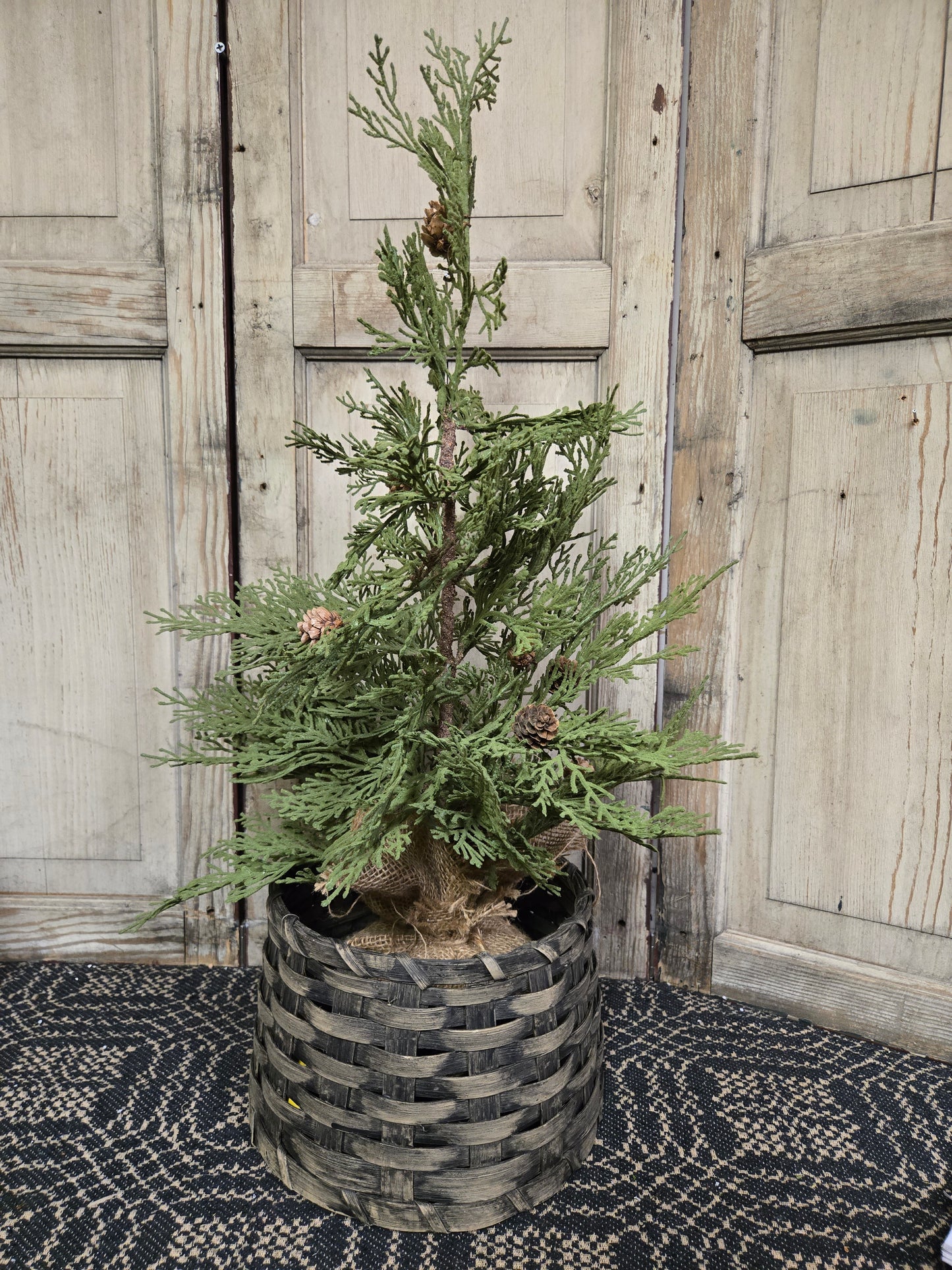 A gray wicker tree collar around a small potted tree, placed on a patterned mat with wooden doors in the background.