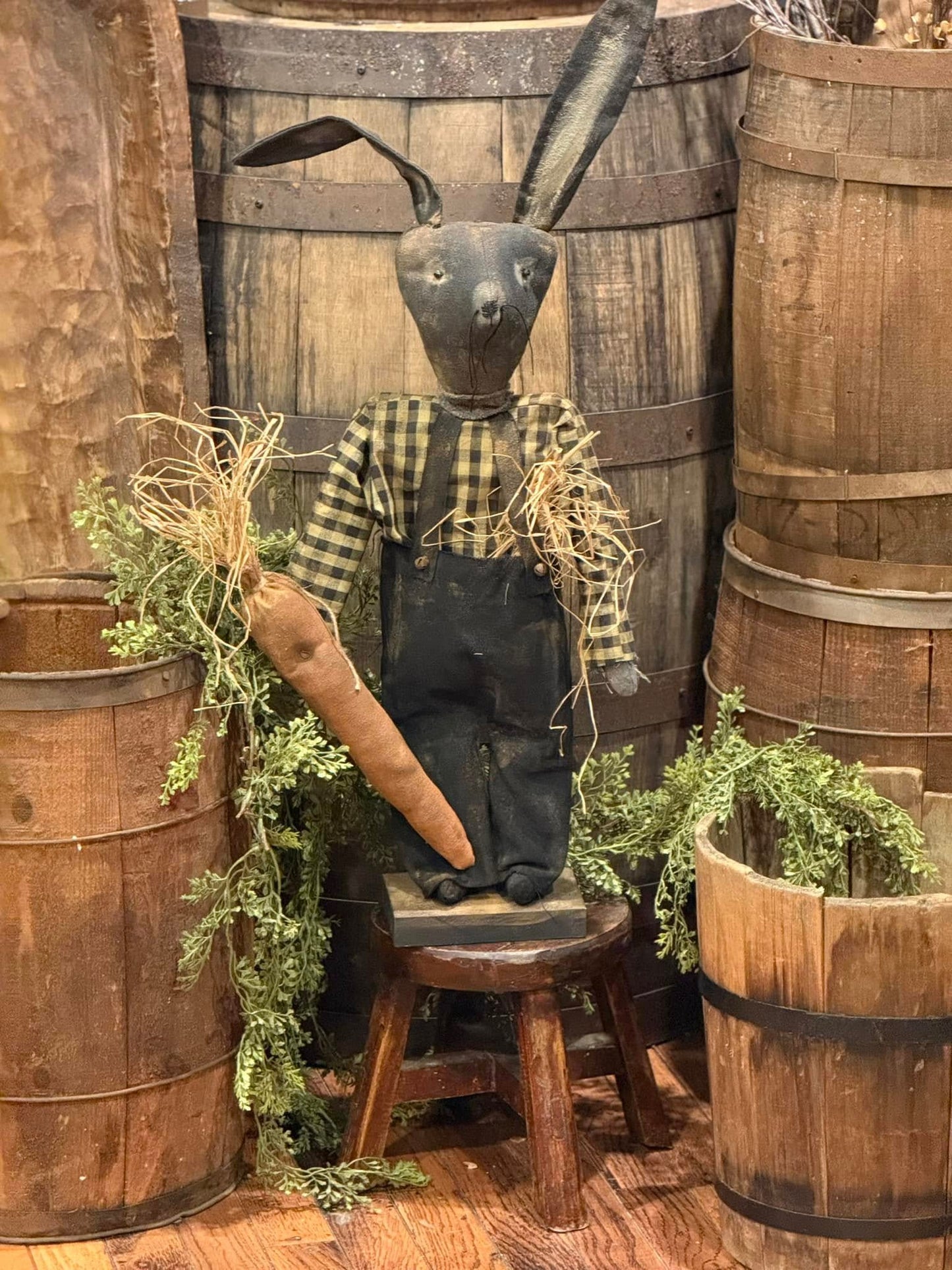 A decorative bunny figure with a black grungy pants and a black and tan checked shirt, sitting in front of wooden barrels.