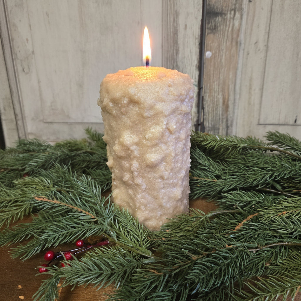 A large pillar candle with a cranberry center, covered in brown sugar and cinnamon, placed on a decorative wreath with green pine branches and red berries.