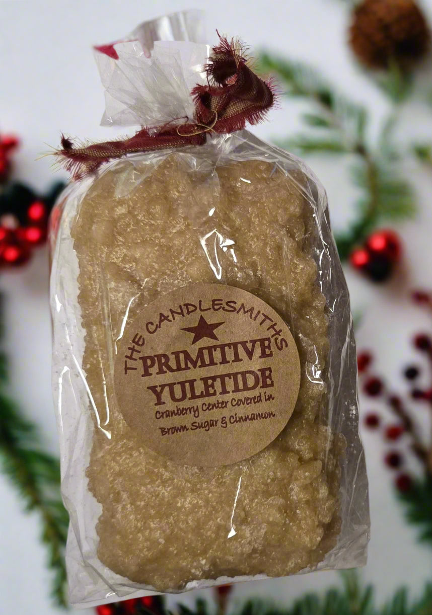 A beige, loaf-shaped candle with a label that reads 'The Primitive Yuletide', packaged in a clear plastic bag with a red ribbon handle.