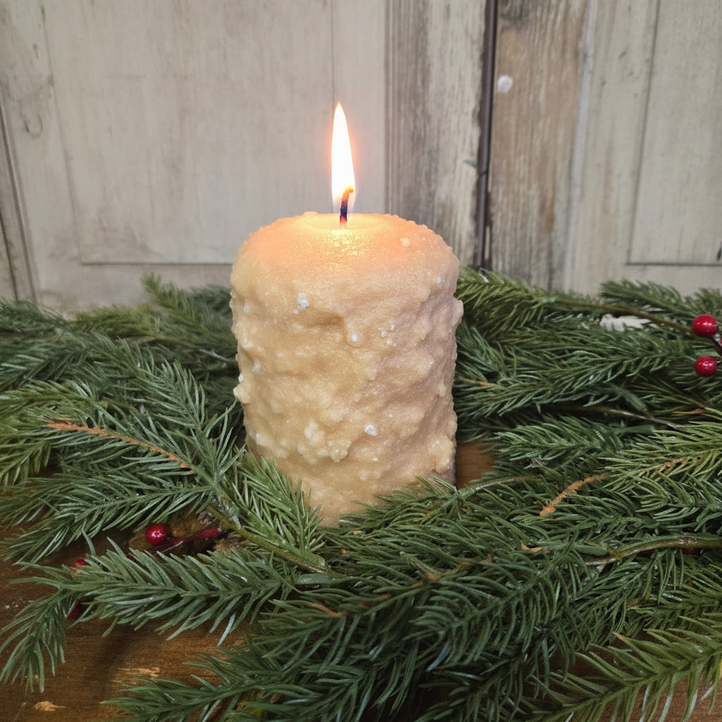 A brown sugar and cinnamon scented pillar candle placed on a wooden surface with green pine branches and red berries around it.