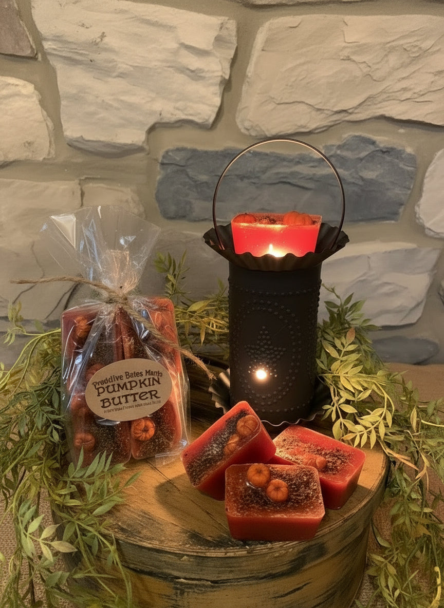A package of orange pumpkin butter wax melts placed in a black metal container, surrounded by green artificial foliage, with a stone wall in the background.