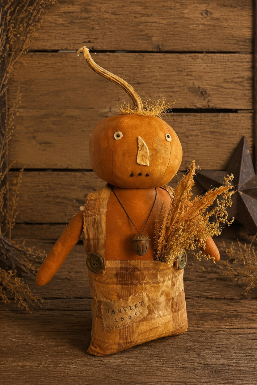 A handmade Primitive Kreation style autumn doll made from a gourd head with a twig nose and stem, wearing a fabric bib over a sack body, holding dried grass, set against a rustic wood plank background with the text 'Primitive Kreations Wholesale'.