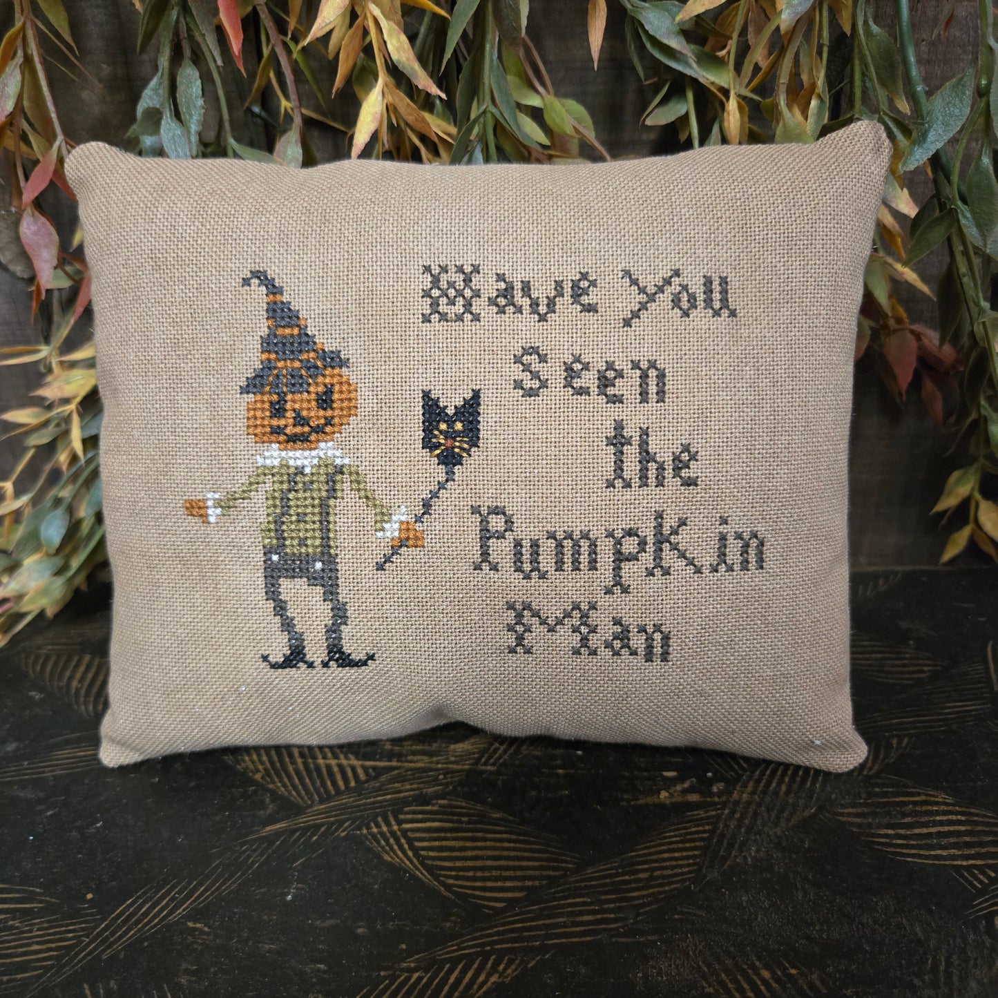 Decorative pillow with cross-stitched design of a pumpkin man and text, surrounded by autumn leaves.