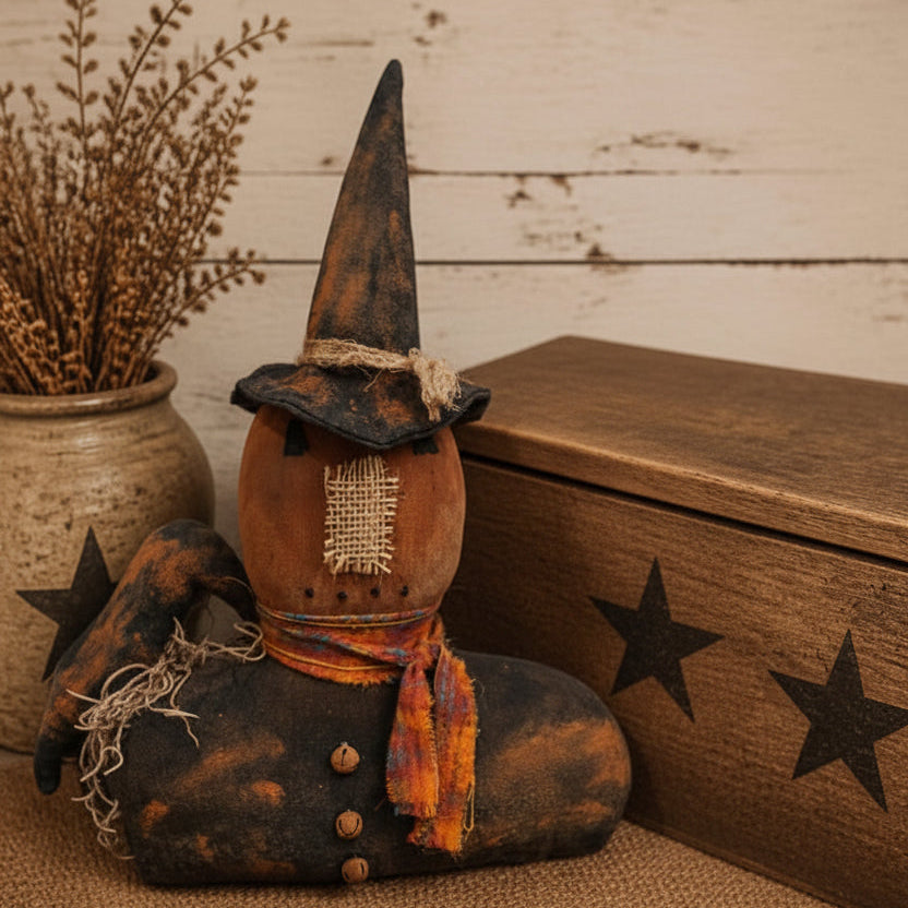 A rustic Halloween decorative figure shaped like a seated figure wearing tall pointed witch hats, a burlap face patch, and an orange plaid scarf, styled with dried grass in a ceramic pot and a wooden box on a burlap runner. The text 'Primitive Kreations Wholesale' appears in the upper right.