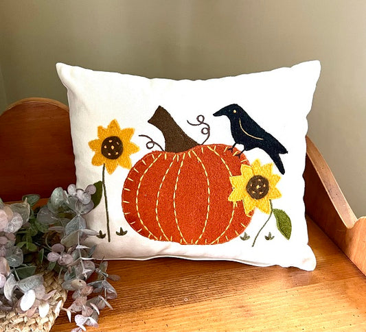 A square-shaped throw pillow with a pumpkin design, featuring sunflowers and a crow on top, placed on a wooden surface.
