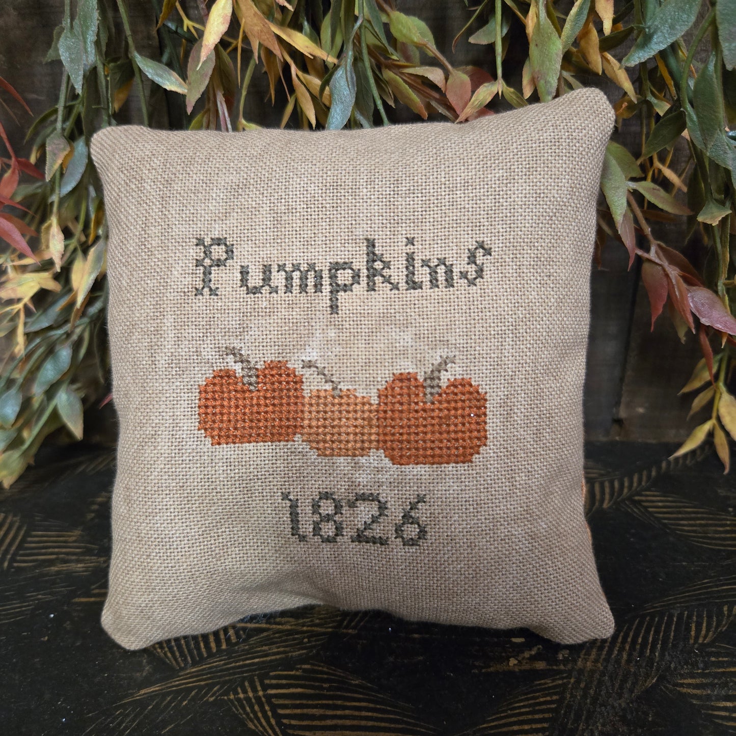 Decorative pillow with cross-stitched pumpkin design and text on a leafy background