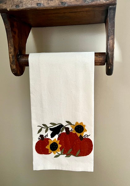 A white hand towel with a decorative pattern featuring pumpkins, sunflowers, and a crow, hanging on a wooden rack.