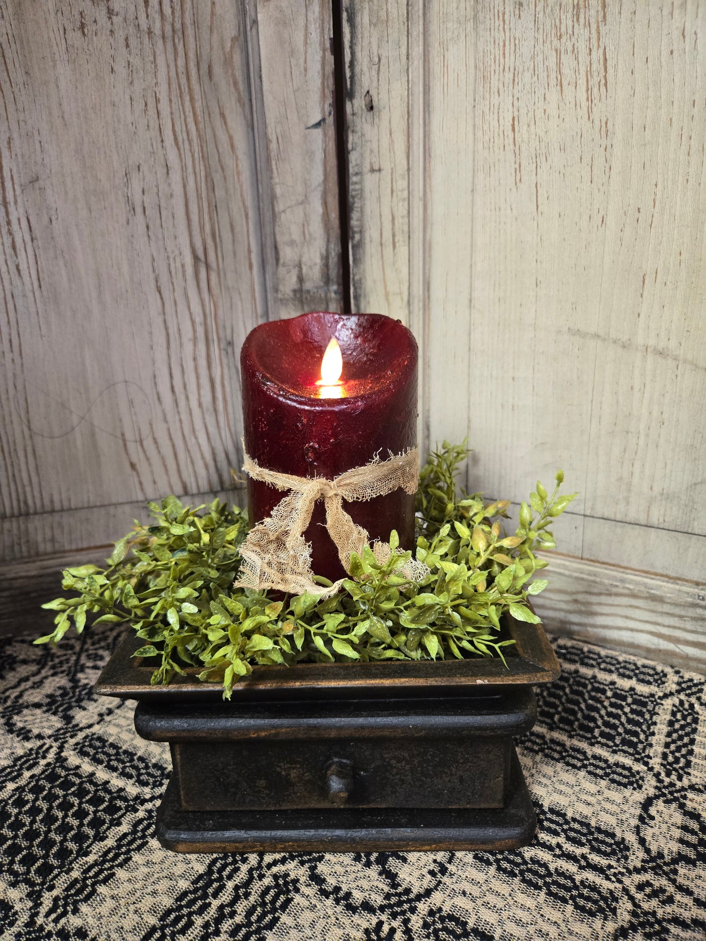 A realistic moving flame 5-inch pillar candle in burgundy color, presented on a wooden surface with a green mossy base inside a brown holder, depicting a realistic appearance.