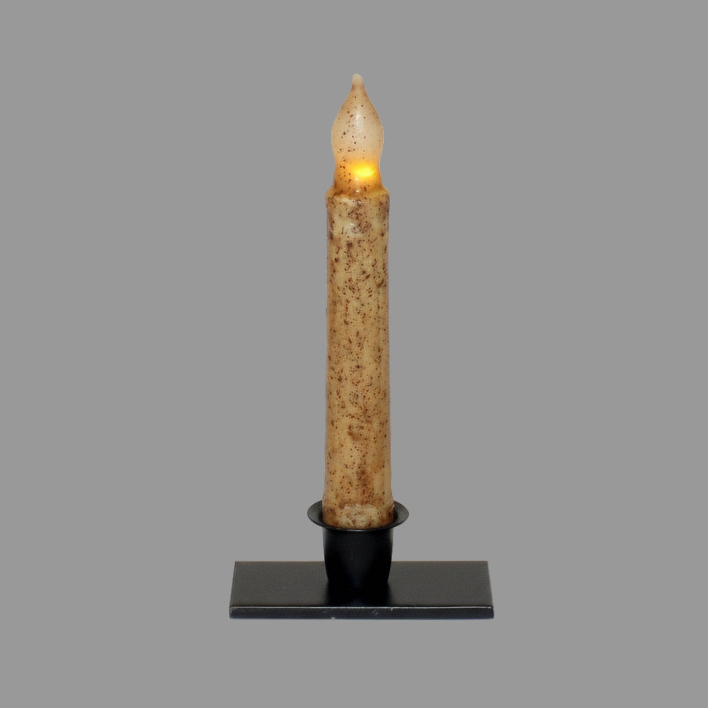 A rectangular candle holder with a tapered design, displaying a candle, placed on a wooden surface with a blurred plant in the background.