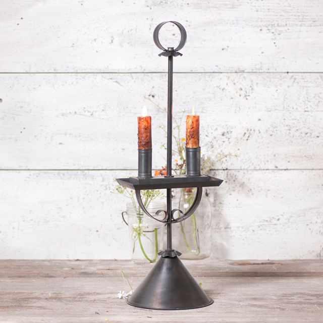 A black metal candle holder with a kettle black finish, featuring two candles, a circular top, and a tapered base, displayed against a rustic backdrop.