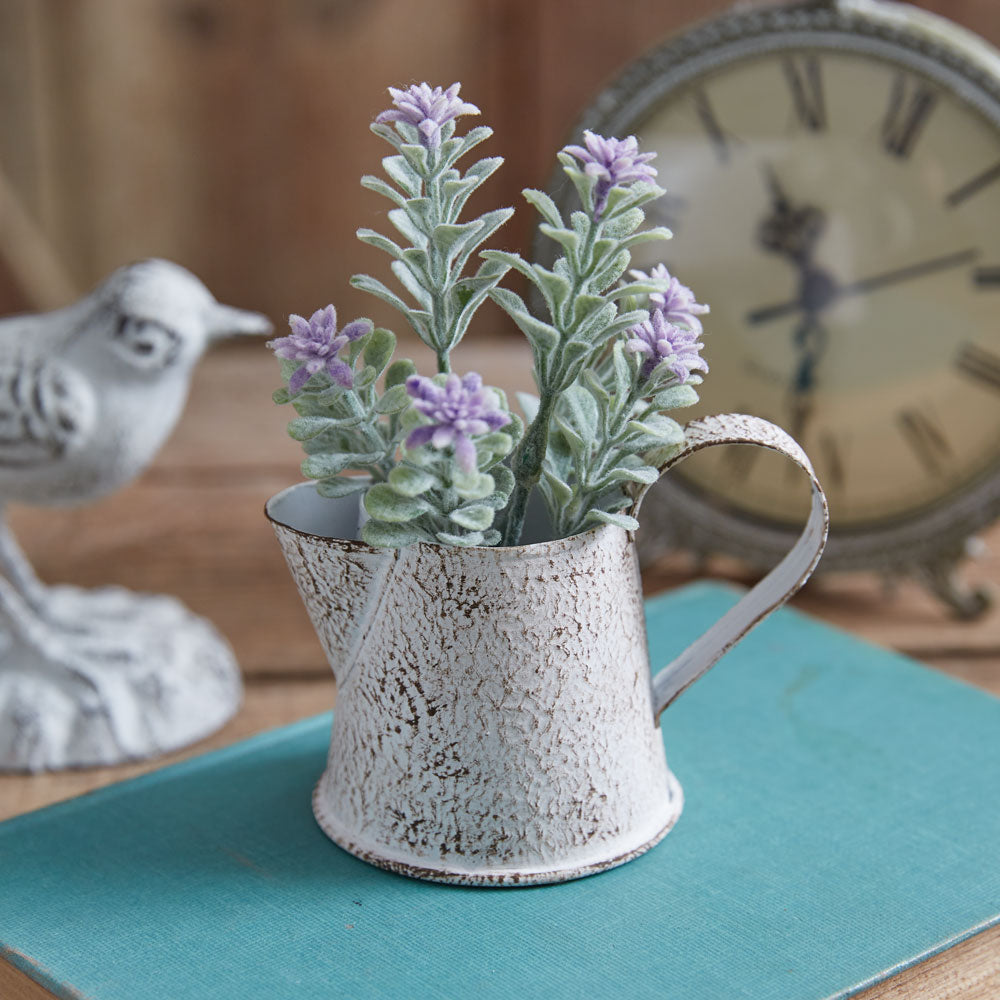 A rustic metal miniature pitcher with a single stem of purple flowers, placed on a teal mat, next to a white birdhouse, a clock, and a bird.