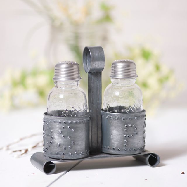 A salt and pepper shaker holder in antique tin finish, with glass shakers, displayed on a white surface with a floral arrangement in the background.