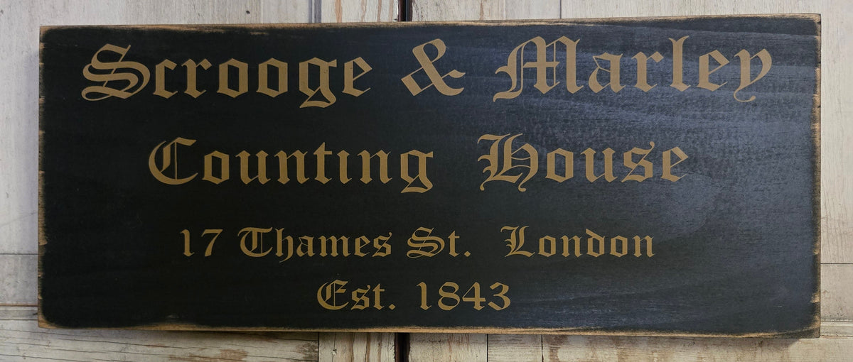 A rustic rectangular wooden sign with a dark black background and gold lettering that reads 'Scrooge & Marley Counting House, 17 Thames St. London, Est.
