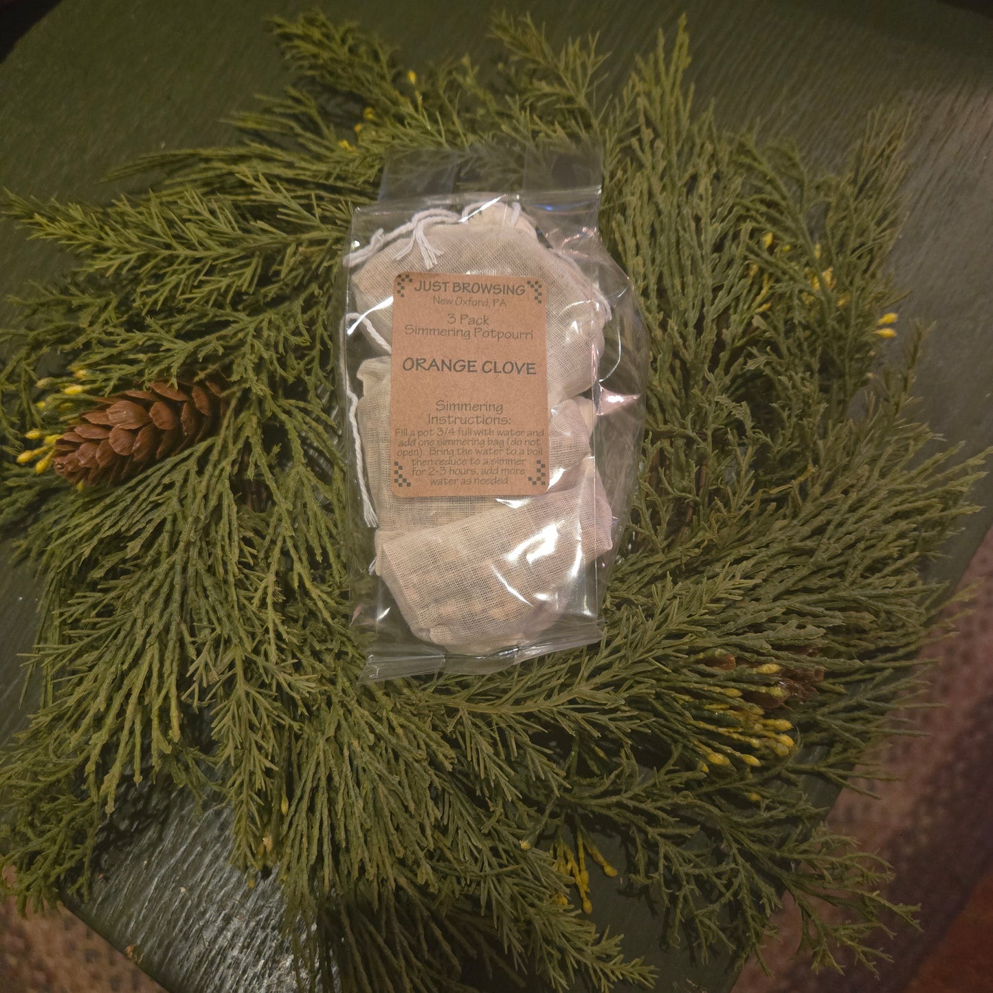 A clear plastic bag filled with orange clove simmering potpourri, placed on a bed of green pine needles and decorative branches.