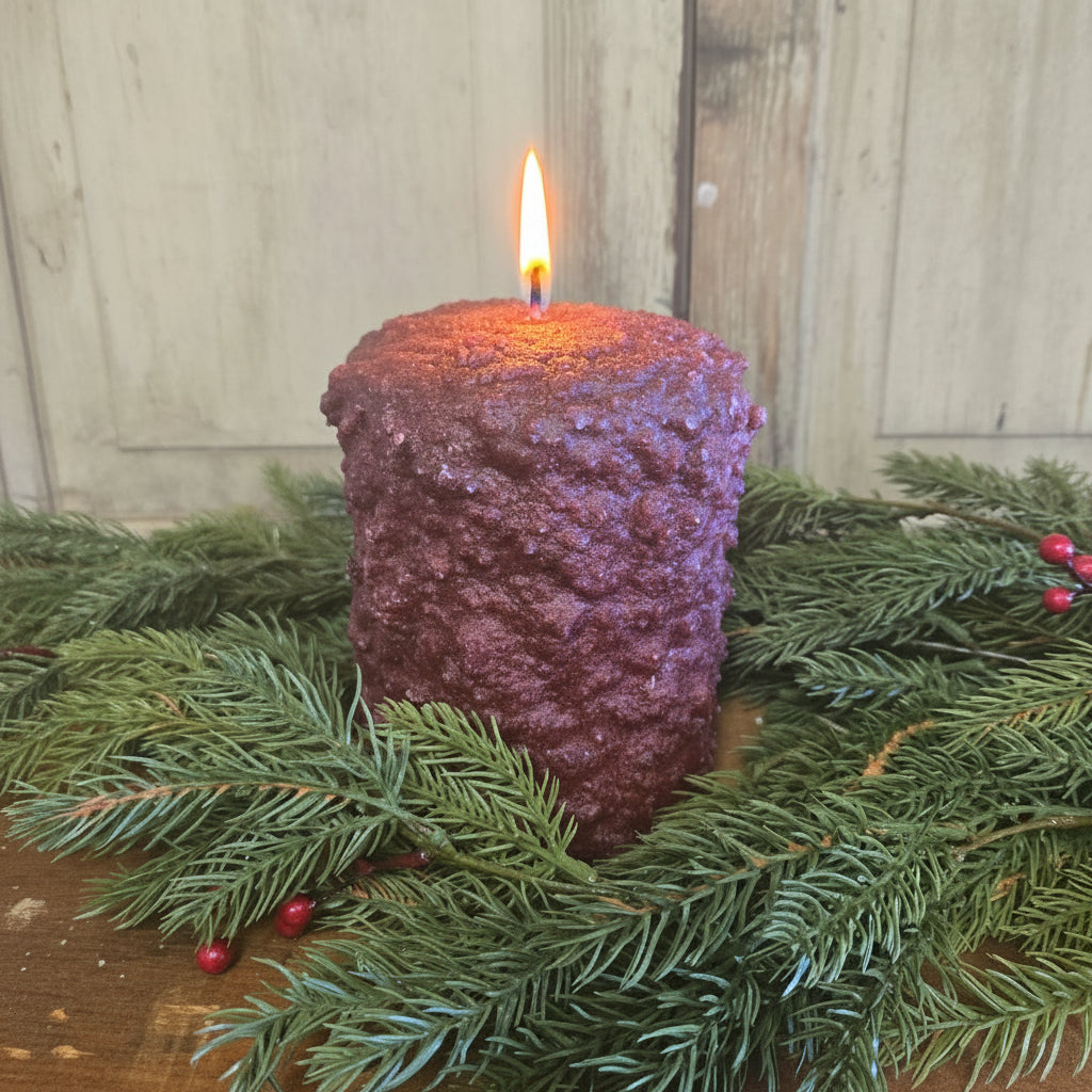 A burgundy colored hearth candle surrounded by greenery against weathered wood