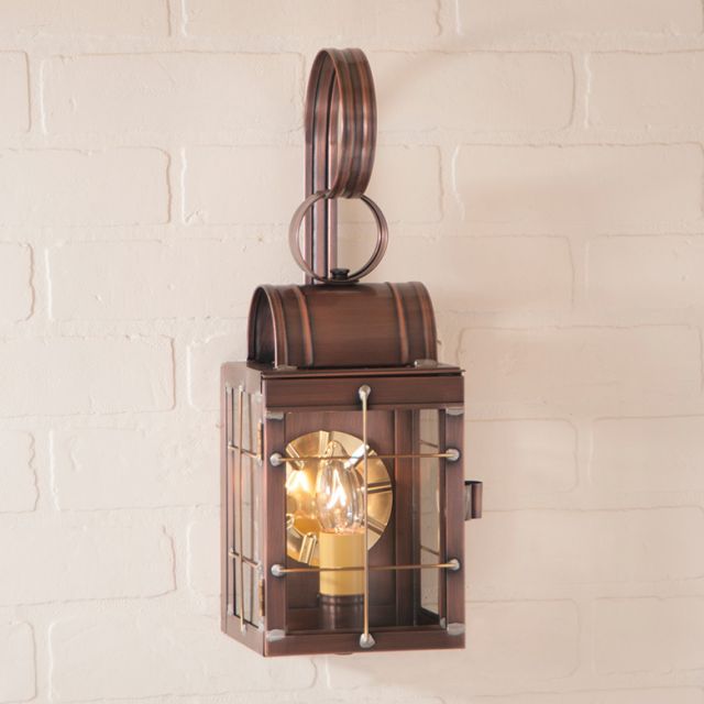 Antique copper wall lantern with a clear glass cover and brass trim, mounted on a white brick wall, emitting a warm light.
