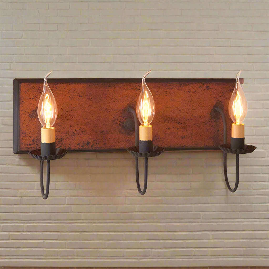 A vintage style vanity light with three bulbs, featuring a distressed pumpkin color over black with black stripe painted finish and textured arms with crimped pans.