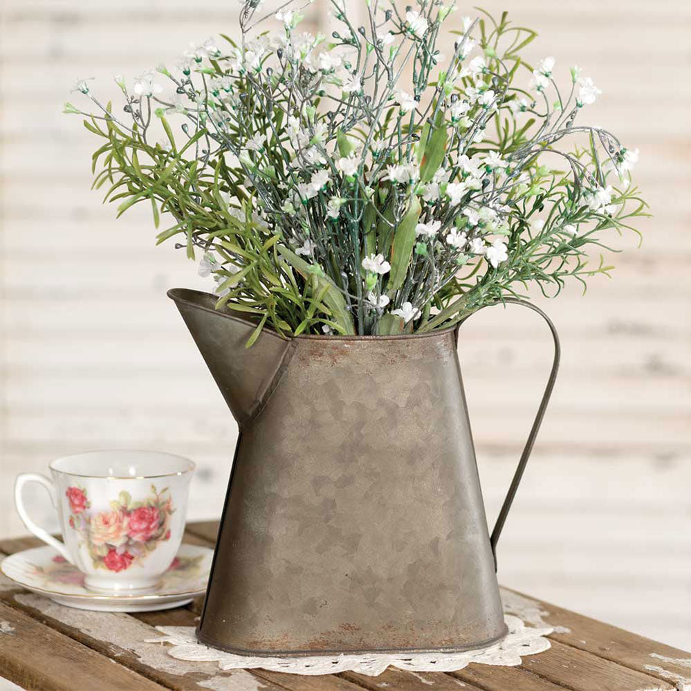 Metal pitcher with artificial flowers on a table next to a teacup and saucer